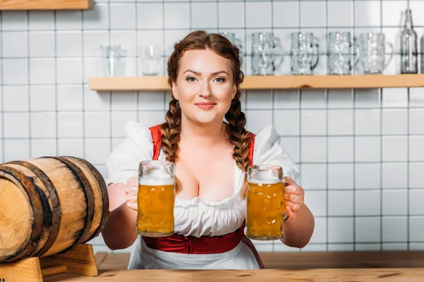 Smiling oktoberfest waitress in traditional bavarian dress giving two mugs of light beer at bar counter — Stock Photo