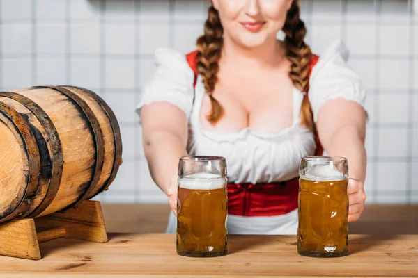 Garçonete oktoberfest em vestido tradicional bávaro colocando canecas de cerveja leve no balcão de bar com barril de cerveja — Fotografia de Stock