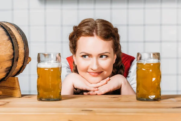 Camarera sonriente oktoberfest en vestido bavariano tradicional sentado entre dos tazas de cerveza ligera en el mostrador del bar - foto de stock