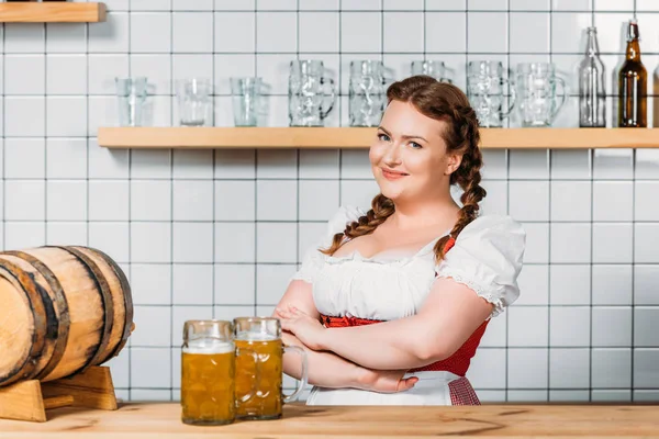 Happy oktoberfest waitress in traditional bavarian dress standing at bar counter with mugs of light beer and beer barrel — Stock Photo