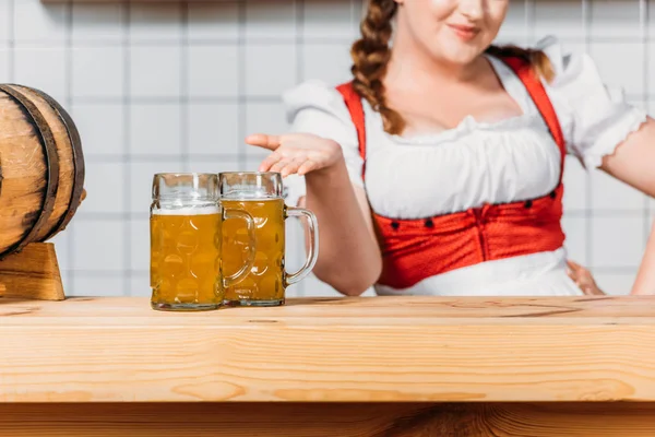 Image recadrée de serveuse oktoberfest en robe bavaroise traditionnelle pointant du doigt des tasses de bière légère sur le comptoir du bar — Photo de stock