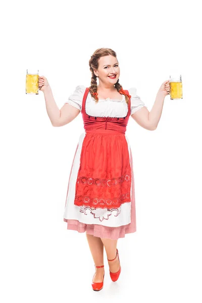 Atractiva camarera oktoberfest en vestido bavariano tradicional sosteniendo tazas de cerveza ligera aislada sobre fondo blanco - foto de stock