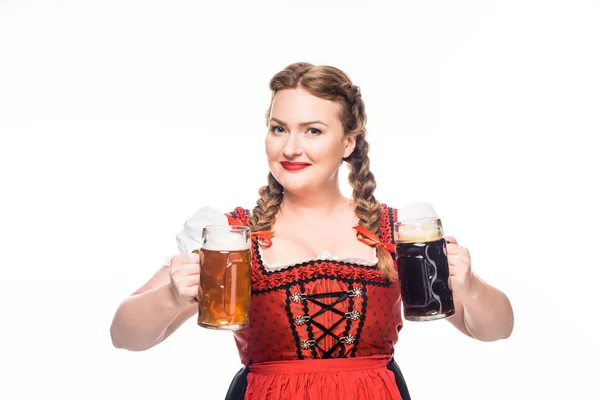 Camarera sonriente oktoberfest en vestido bavariano tradicional mostrando tazas con cerveza clara y oscura aislada sobre fondo blanco - foto de stock