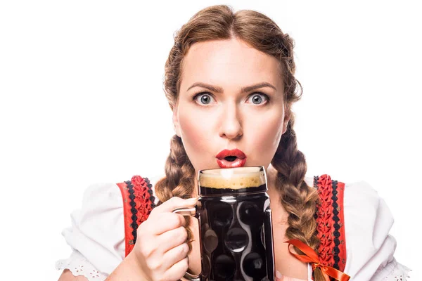 Shocked oktoberfest waitress in traditional bavarian dress holding mug of dark beer isolated on white background — Stock Photo