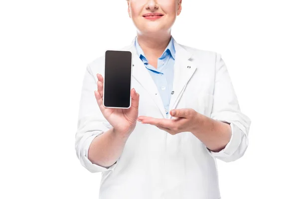 Обрезанное изображение женщины-врача, указывающей на смартфон с пустым экраном, изолированным на белом фоне — стоковое фото
