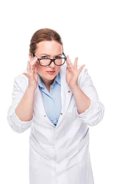 Confident female doctor adjusting eyeglasses and looking at camera isolated on white background — Stock Photo