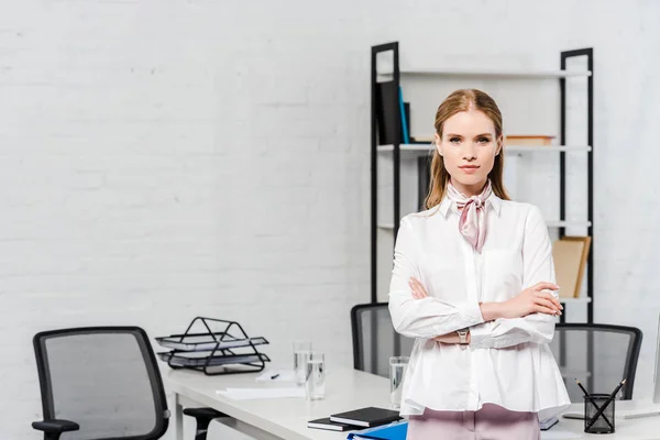 Beautiful young businesswoman with crossed arms looking at camera at modern office — Stock Photo