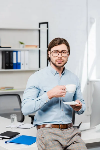 Joven hombre de negocios guapo con taza de café mirando a la cámara en la oficina moderna - foto de stock