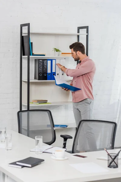 Schöner junger Geschäftsmann nimmt Ordner aus Bücherregal in modernem Büro — Stockfoto