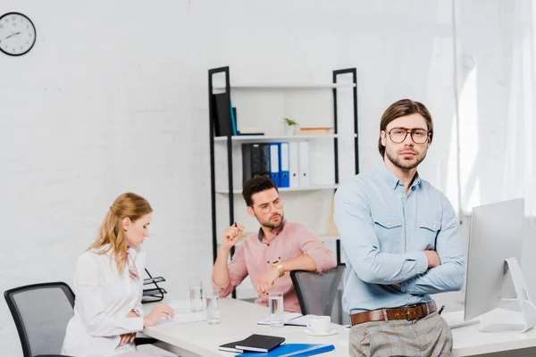 Schöner junger Geschäftsmann mit verschränkten Armen, der in die Kamera blickt und sich im modernen Büro auf den Tisch zurücklehnt, während Kollegen im Hintergrund zusammenarbeiten — Stock Photo