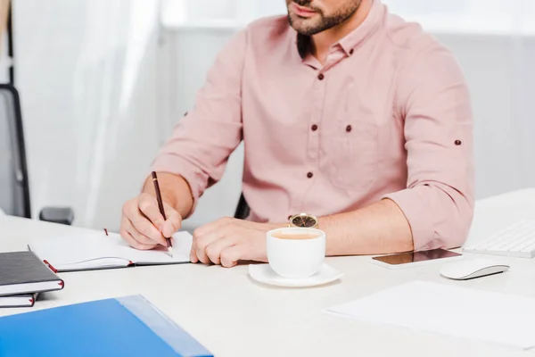 Обрезанный снимок молодого бизнесмена с чашкой кофе делая заметки на рабочем месте — стоковое фото