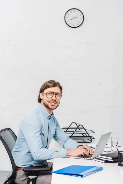 Sorridente jovem empresário trabalhando com laptop no escritório moderno e olhando para a câmera — Fotografia de Stock