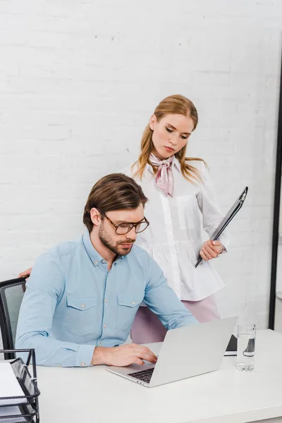 Selbstbewusste junge Managerin, die neben dem Chef steht, während er im modernen Büro Laptop benutzt — Stockfoto