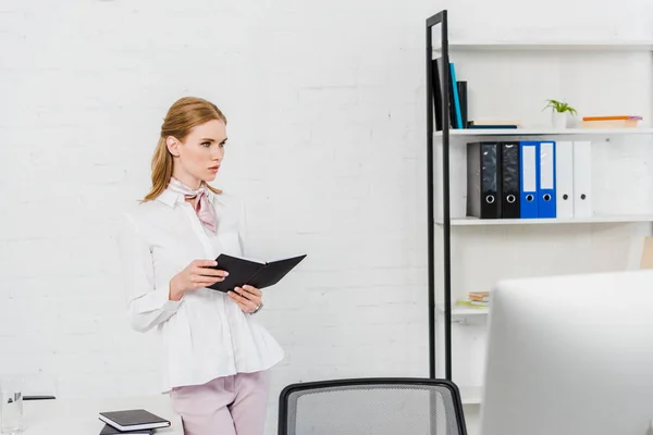 Ernsthafte junge Geschäftsfrau mit Buch steht im modernen Büro und schaut weg — Stockfoto