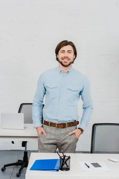 Handsome young businessman looking at camera with hands in pockets at modern office — Stock Photo