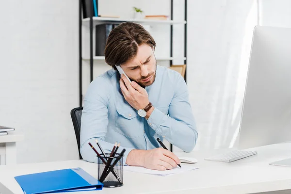 Giovane uomo d'affari fiducioso che parla per telefono e prende appunti sul posto di lavoro — Foto stock