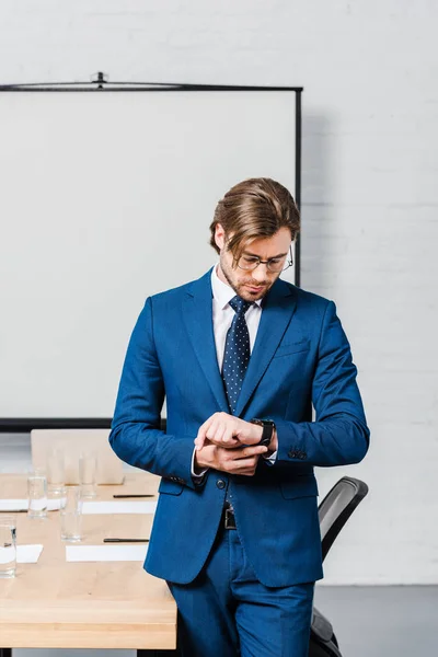 Apuesto joven hombre de negocios mirando reloj de pulsera en la sala de conferencias con tablero de presentación en blanco - foto de stock