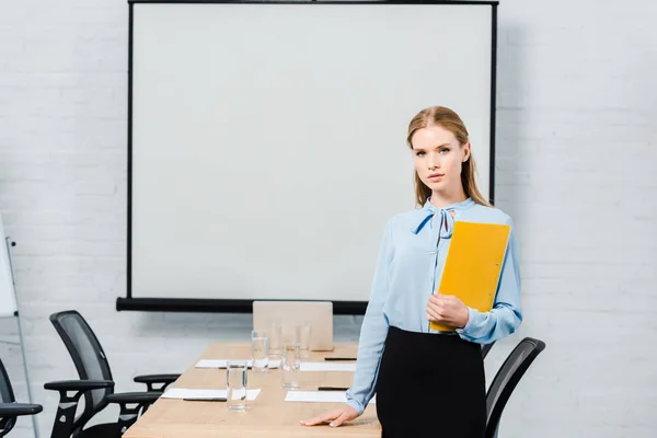 Selbstbewusste junge Geschäftsfrau mit Mappe blickt in Konferenzsaal mit leerer Präsentationstafel in die Kamera — Stockfoto