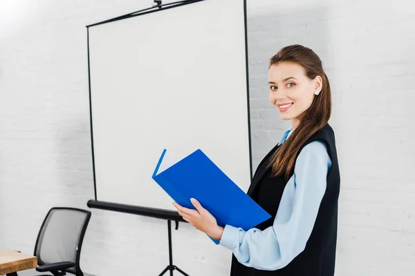 Smiling young businesswoman with folder of documents looking at camera in front of blank presentation board — Stock Photo