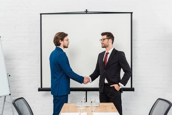 Beaux hommes d'affaires serrant la main devant le tableau de présentation au bureau moderne — Photo de stock