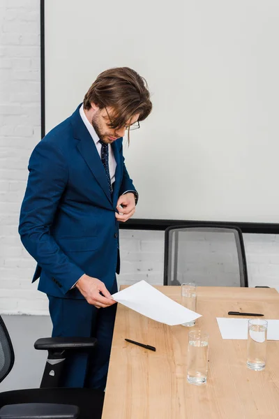 Handsome young businessman reading documents and buttoning jacket at modern office — Stock Photo