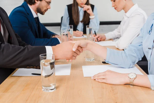 Recortado disparo de socios de negocios estrechando la mano durante la reunión en la oficina moderna - foto de stock