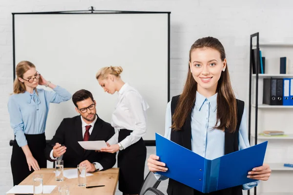 Улыбающаяся молодая деловая женщина с документами, стоящими в современном офисе, с коллегами, беседующими на заднем плане — стоковое фото