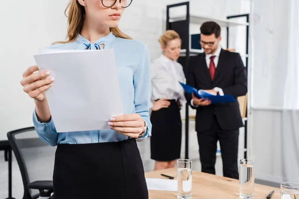 Plan recadré de jeune femme d'affaires avec des documents debout au bureau moderne avec des collègues sur fond — Photo de stock