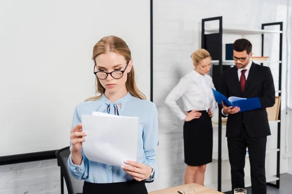 Giovane donna d'affari attraente con documenti in piedi a ufficio moderno con colleghi su sfondo — Foto stock