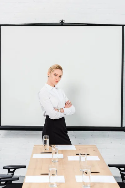 Hermosa joven mujer de negocios con brazos cruzados delante del tablero de presentación en blanco en la sala de conferencias - foto de stock