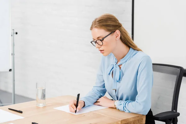 Bela jovem empresária escrevendo em papel em branco no escritório moderno — Fotografia de Stock