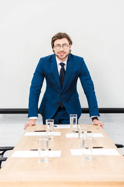 Lächelnder junger Geschäftsmann, der in die Kamera blickt, während er sich im Konferenzsaal auf den Tisch lehnt und in die Kamera blickt — Stockfoto