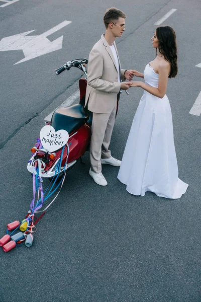 Pareja de boda de la mano en la carretera cerca de scooter con signo de corazón 