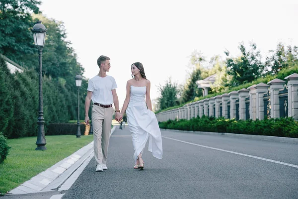 Mariée en robe de mariée et marié tenant la main et marchant sur la route — Photo de stock