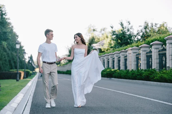 Attractive bride and handsome groom holding hands and walking on road — Stock Photo