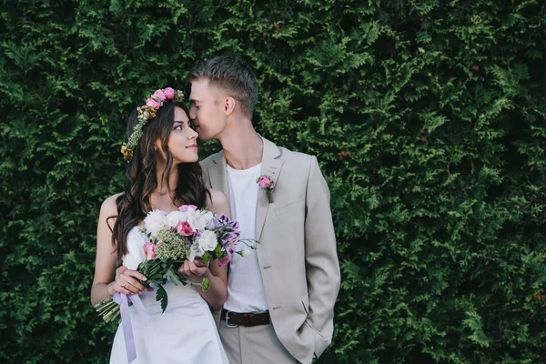 Bräutigam küsst und umarmt schöne Braut in traditionellem Kleid mit Kranz und Brautstrauß — Stockfoto