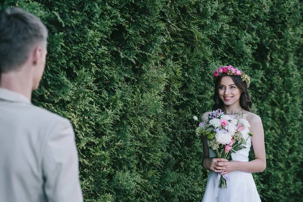 Foyer sélectif de mariée attrayante heureuse en couronne tenant bouquet de mariage et regardant marié — Photo de stock