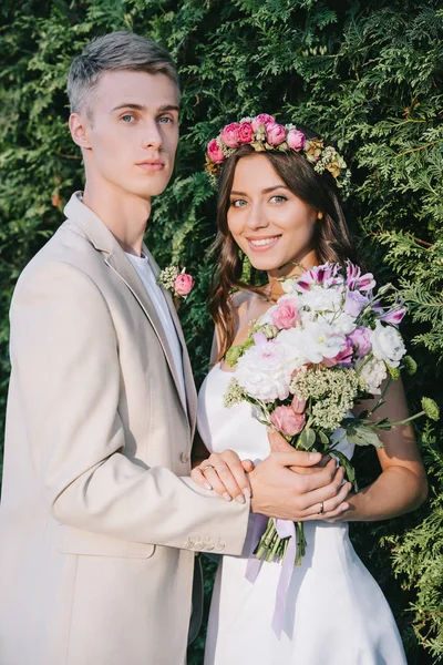 Элегантная свадебная пара с цветочным букетом, смотрящая в камеру — стоковое фото