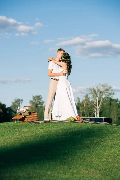 Wedding couple embracing and kissing on green lawn with picnic — Stock Photo