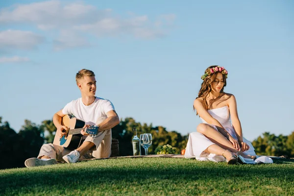 Groom playing guitar for beautiful bride during romantic picnic with wine and fruits — Stock Photo