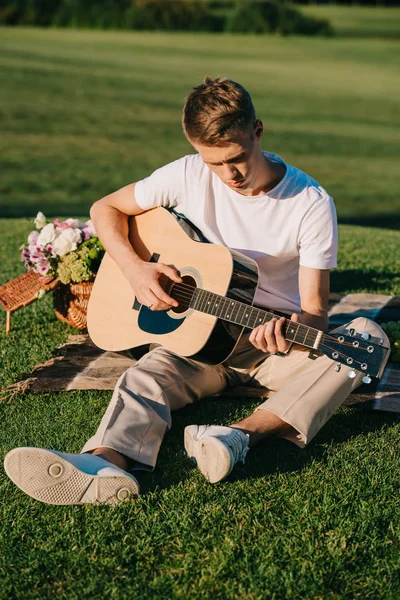 Молодий чоловік грає на акустичній гітарі під час пікніка на зеленому газоні — стокове фото