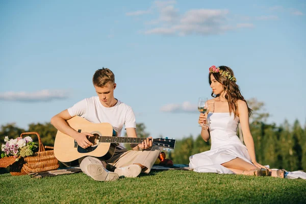 Novio guapo tocando en la guitarra acústica para novia con vino durante el picnic romántico — Stock Photo