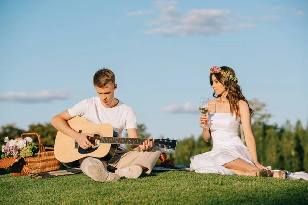 Handsome groom playing on acoustic guitar for bride with wine during romantic picnic — Stock Photo