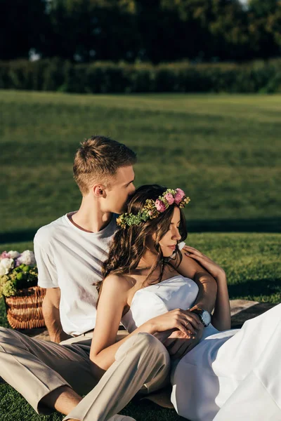 Guapo novio abrazando hermosa novia en vestido de novia y corona en el picnic - foto de stock