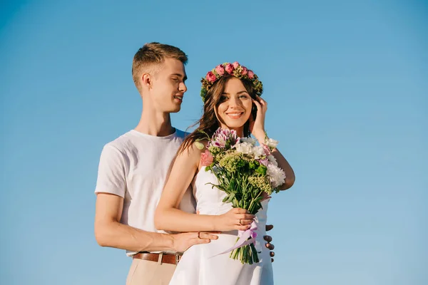 Schönes Hochzeitspaar hält Blumenstrauß in der Hand und posiert vor blauem Himmel — Stockfoto