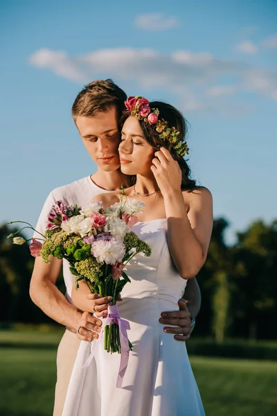 Novio abrazando a su atractiva novia en corona de flores con ramo de bodas - foto de stock
