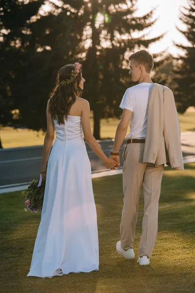 Vue arrière de la mariée attrayante et beau marié tenant la main à l'extérieur — Photo de stock