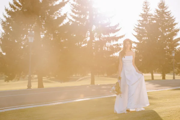 Attractive bride in white dress holding wedding bouquet, outdoors with back light — Stock Photo