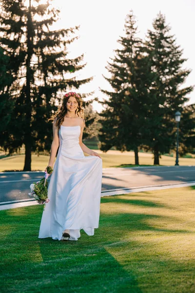 Красивая счастливая молодая невеста, держащая свадебный букет и улыбающаяся перед камерой в парке — стоковое фото