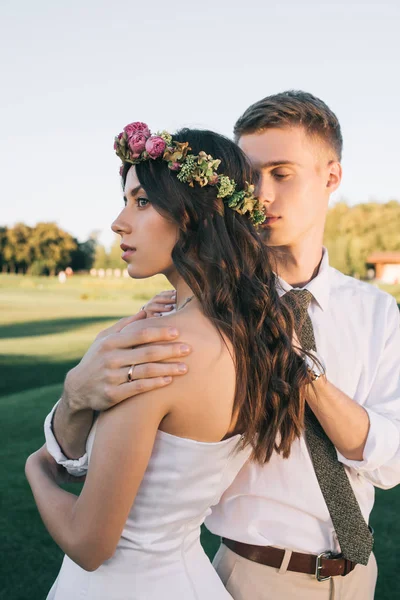 Young groom embracing beautiful young bride in floral wreath at park — Stock Photo